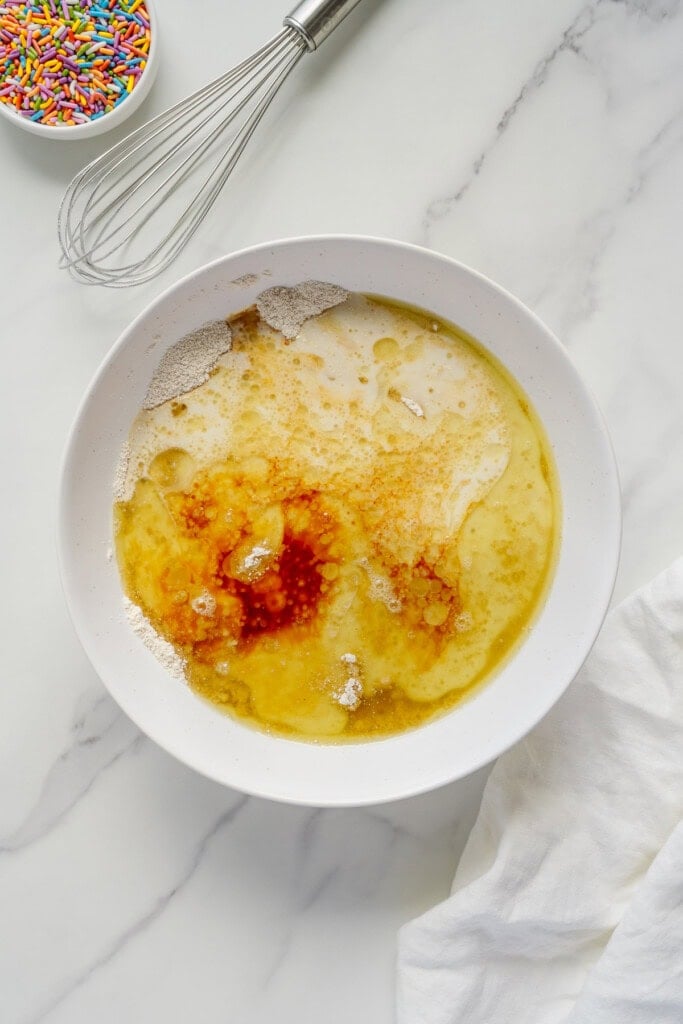 A white mixing bowl with melted butter, vanilla, and other ingredients sits on a marble surface next to a whisk, a bowl of colorful sprinkles, and a white cloth.
