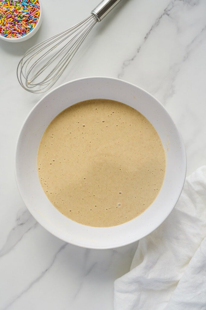 A white bowl filled with pancake batter sits on a marble surface next to a whisk, a small bowl of colorful sprinkles, and a white cloth.