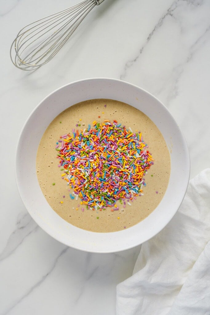A white bowl filled with cake batter and topped with colorful rainbow sprinkles sits on a marble surface, with a metal whisk and a white cloth nearby.