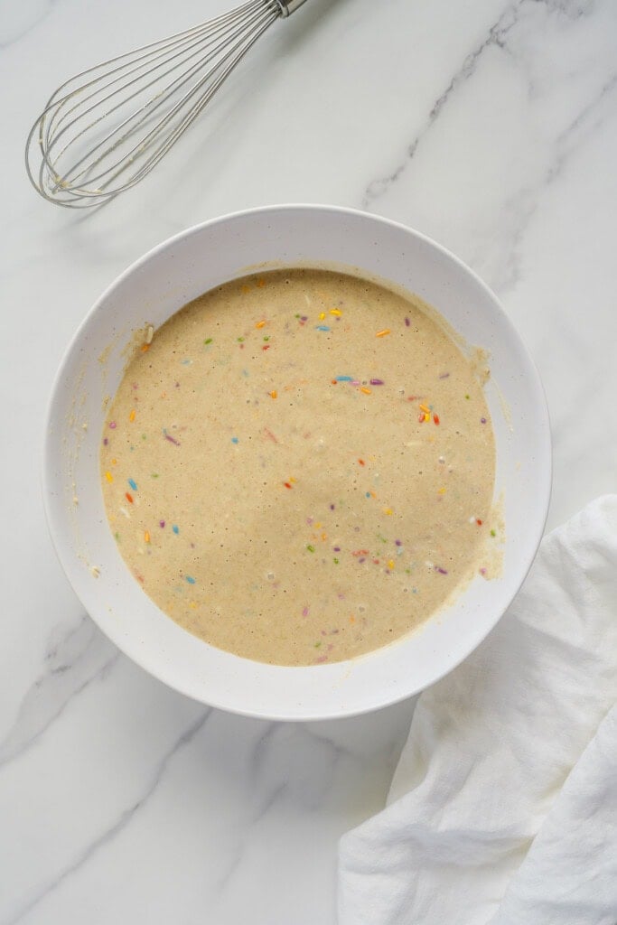 A white bowl filled with cake batter mixed with colorful sprinkles sits on a marble surface, next to a wire whisk and a white cloth.