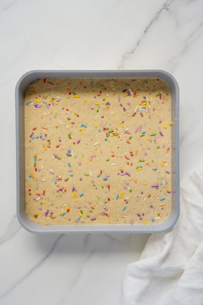 A square baking pan filled with unbaked cake batter topped with colorful rainbow sprinkles, set on a white marble surface with part of a white cloth visible on the side.