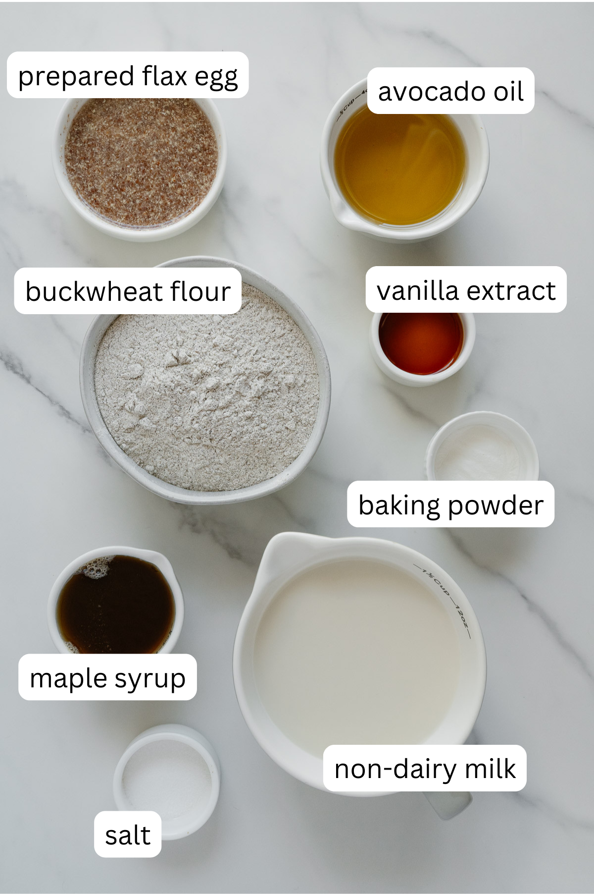 Top-down view of labeled bowls and cups containing prepared flax egg, avocado oil, buckwheat flour, vanilla extract, baking powder, maple syrup, non-dairy milk, and salt on a white marble surface.