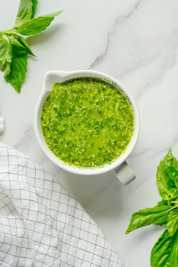 A white pitcher filled with green pesto sauce sits on a marble surface, surrounded by fresh basil leaves and a white kitchen towel with black grid lines.