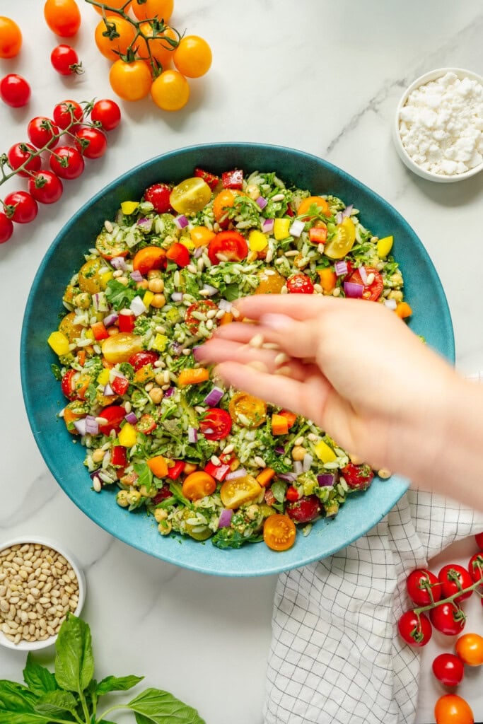 A hand sprinkles pine nuts over a colorful chopped salad in a blue bowl, surrounded by cherry tomatoes, feta cheese, fresh basil, and a white dish towel on a marble countertop.