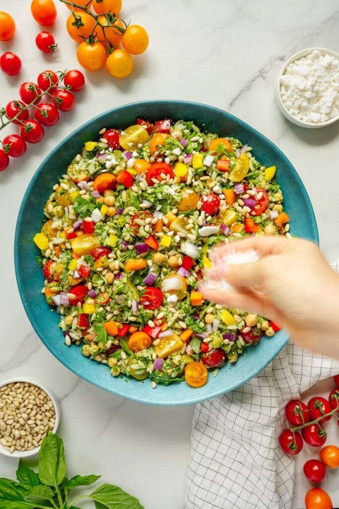 A hand sprinkles cheese over a colorful salad with cherry tomatoes, chickpeas, red onion, and herbs in a blue bowl. Surrounding the bowl are basil, cherry tomatoes, pine nuts, cheese, and a white towel on a marble surface.