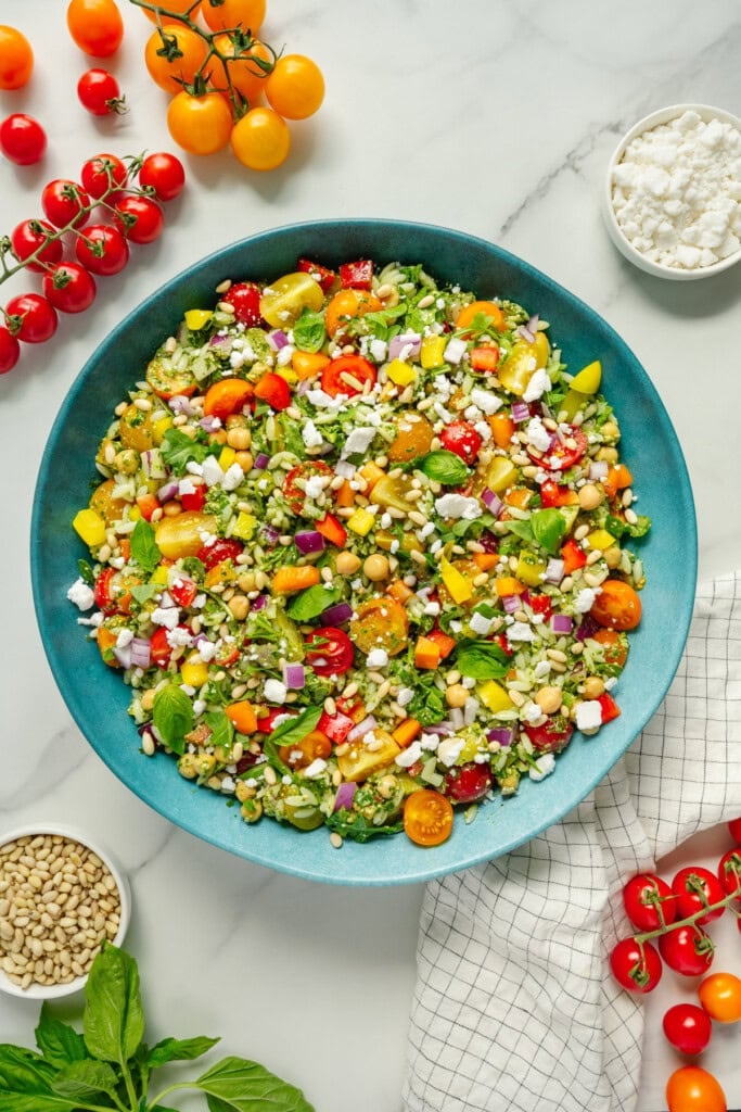 A large blue bowl filled with colorful chopped salad, including tomatoes, corn, red onion, feta cheese, bell peppers, and herbs. Surrounding the bowl are cherry tomatoes, basil, pine nuts, and a small dish of feta.