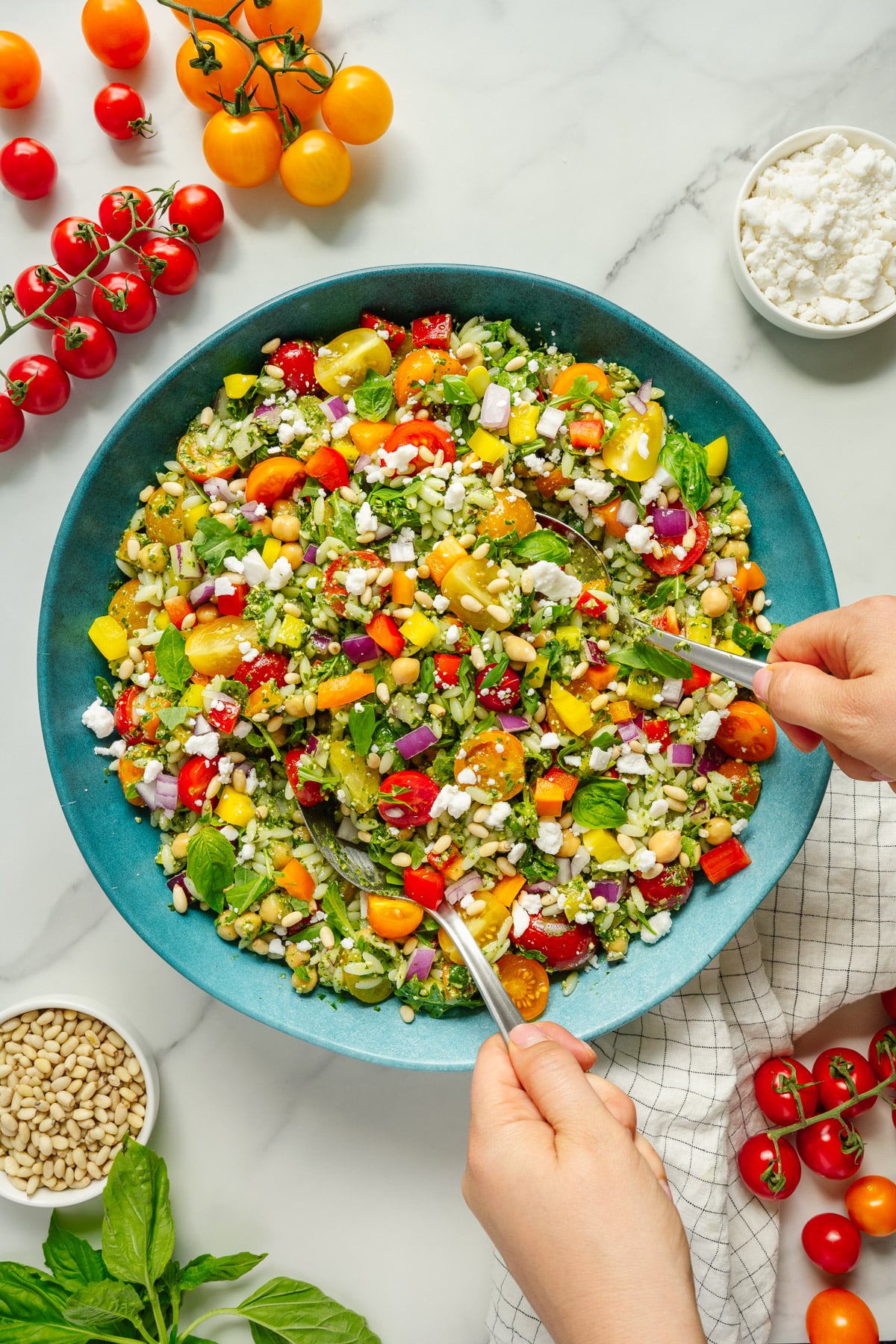 A person tosses a colorful salad with tomatoes, bell peppers, red onions, greens, feta cheese, and grains in a blue bowl, surrounded by fresh vegetables, pine nuts, basil, and a small bowl of feta on a marble surface.