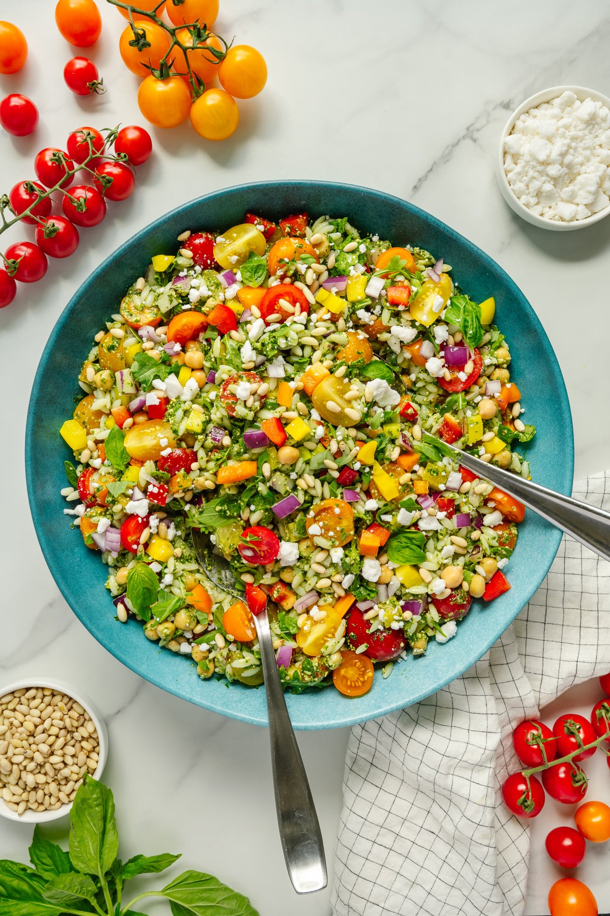 A large blue bowl filled with colorful chopped salad, including cherry tomatoes, feta cheese, cucumbers, red onion, and herbs, with two serving spoons. Surrounding the bowl are bowls of feta, pine nuts, fresh basil, and cherry tomatoes.