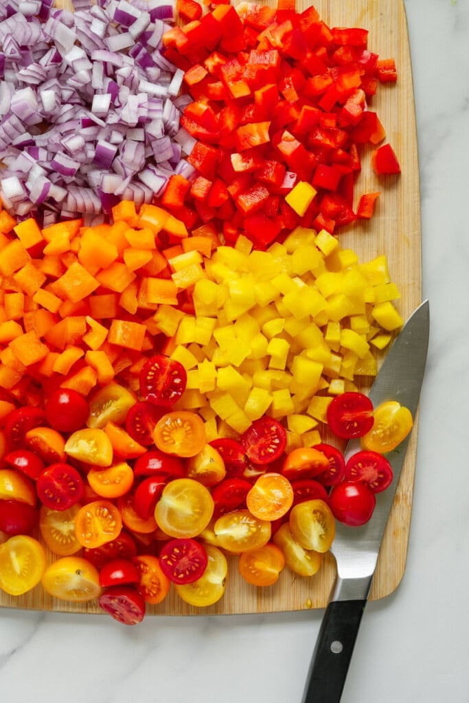 A cutting board with diced red onion, red bell pepper, orange bell pepper, yellow bell pepper, and halved red and yellow cherry tomatoes, next to a knife on a marble surface.