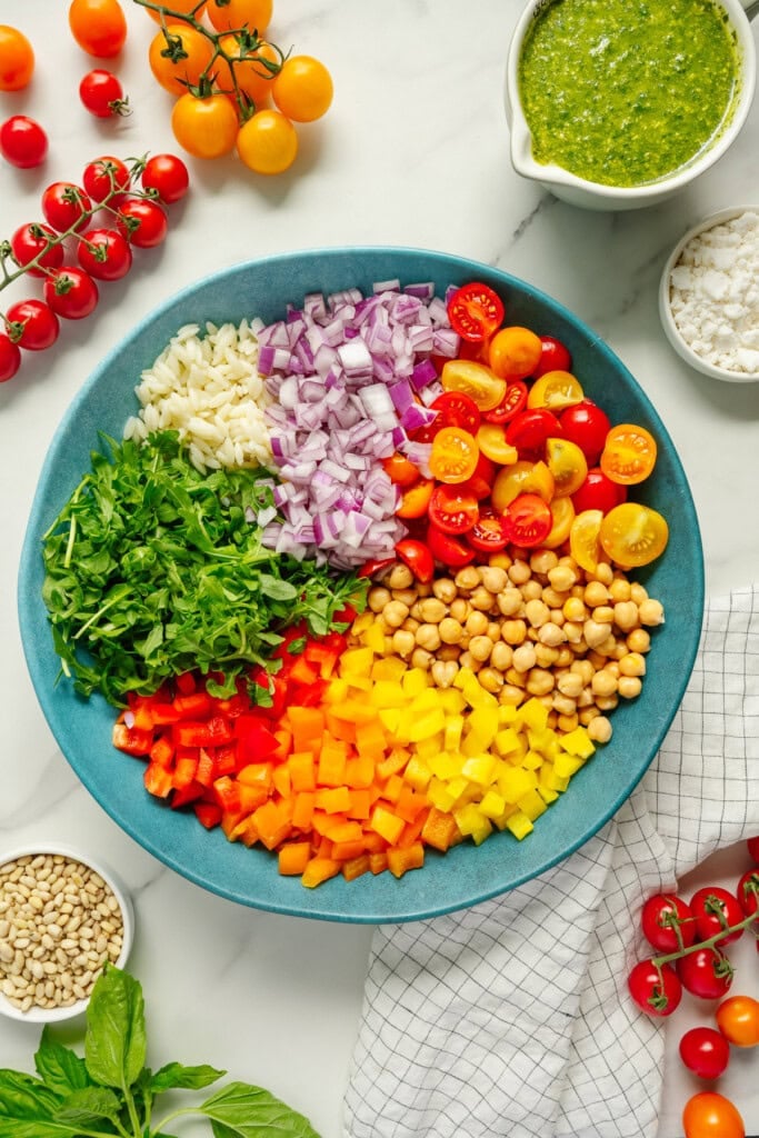 A large blue bowl filled with colorful chopped vegetables, chickpeas, and arugula arranged in sections. Surrounding the bowl are cherry tomatoes, pine nuts, a bowl of green pesto, basil, and a cloth napkin.