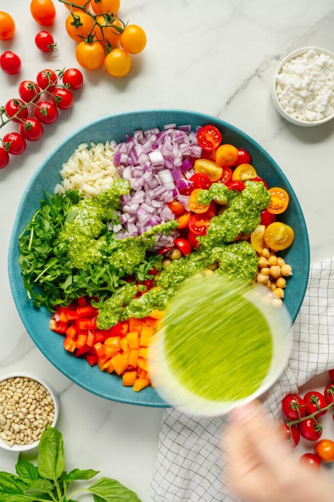 A large blue bowl filled with chopped vegetables, chickpeas, cherry tomatoes, arugula, onions, and pesto. A hand pours green dressing over the salad. Ingredients like tomatoes, feta cheese, pine nuts, and basil surround the bowl.