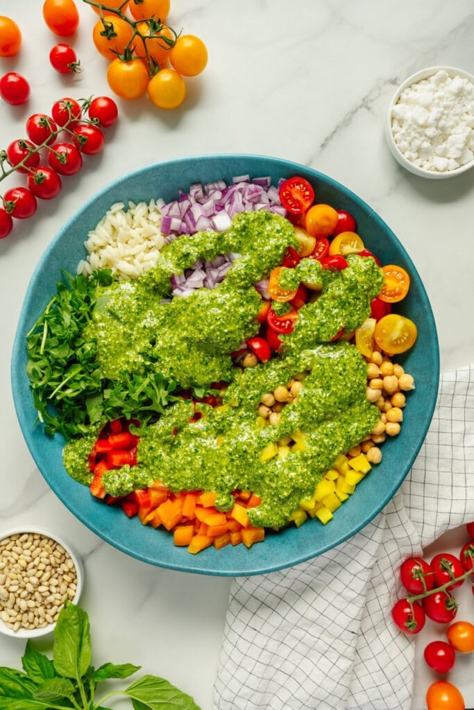 A colorful salad with diced red onion, cherry tomatoes, chickpeas, red and yellow bell peppers, arugula, and pesto dressing in a blue bowl, surrounded by fresh tomatoes, pine nuts, cheese, and basil on a white surface.