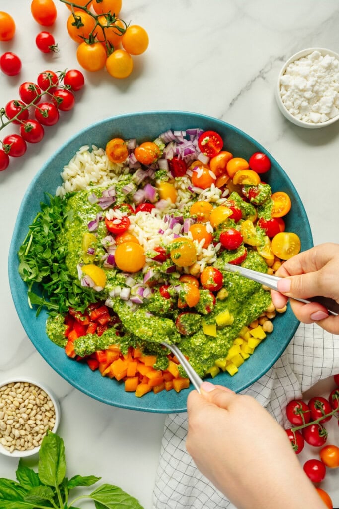 A colorful salad with cherry tomatoes, chopped onions, bell peppers, chickpeas, greens, pesto, and cheese in a blue bowl. Two hands mix the ingredients with utensils. Tomatoes, pine nuts, and cheese are nearby on a marble surface.