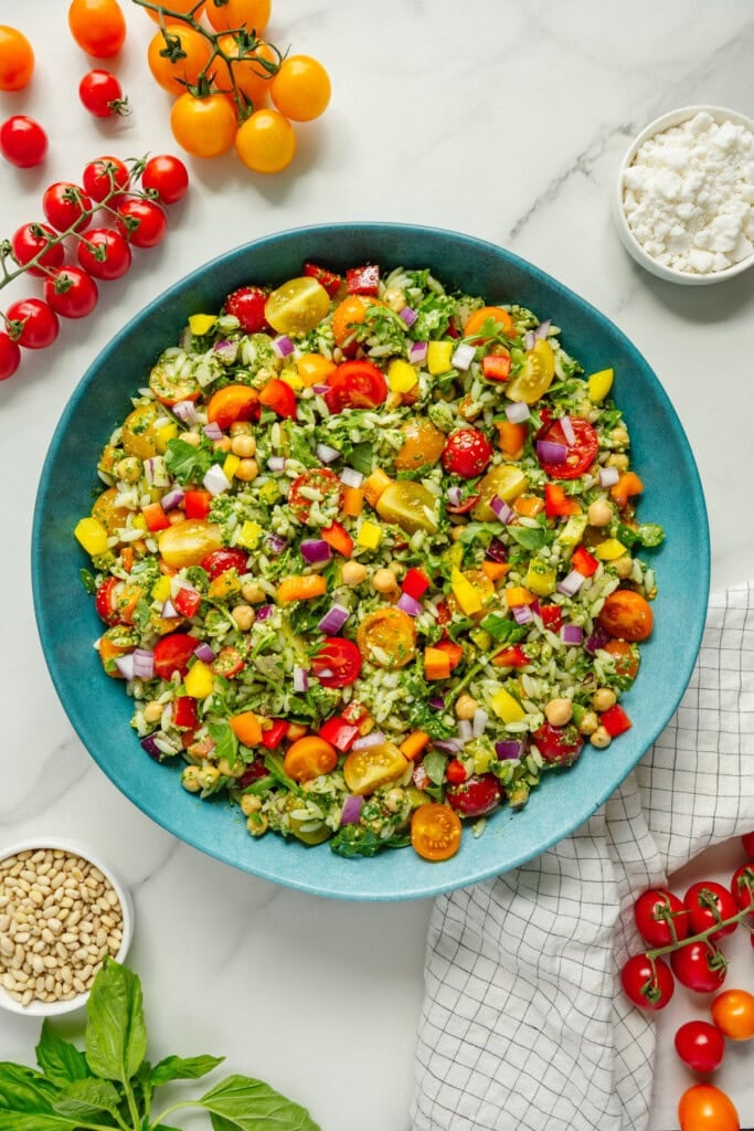 Colorful chopped salad with cherry tomatoes, cucumbers, bell peppers, red onion, chickpeas, and feta in a blue bowl. Surrounding the bowl are cherry tomatoes, basil, pine nuts, and a white towel on a marble surface.