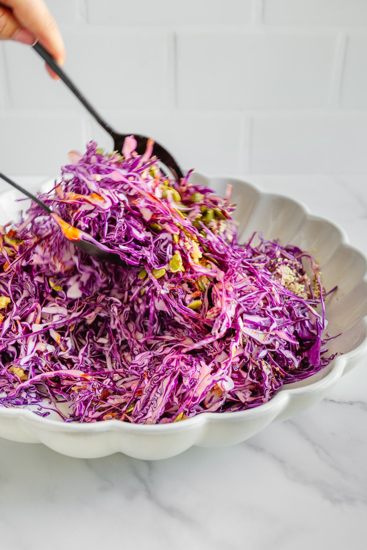 A hand uses black salad tongs to mix shredded purple cabbage and vegetables in a white scalloped bowl on a marble surface.