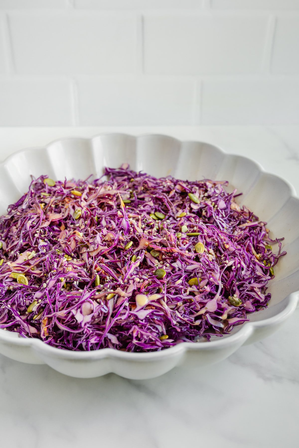 A white scalloped bowl filled with finely shredded purple cabbage salad, garnished with seeds, sits on a white marble surface against a white tiled background.