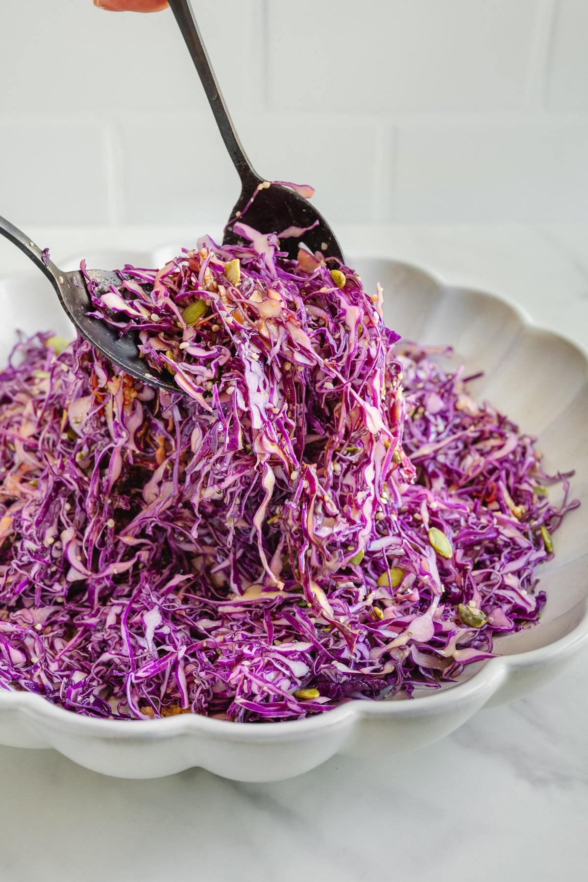 A hand holds tongs mixing shredded purple cabbage salad with seeds in a scalloped white bowl on a white surface.