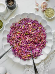Large white scalloped bowl filled with shredded purple cabbage slaw with seeds, placed on a light surface with garlic cloves, spices, and utensils around. Two serving spoons rest in the bowl.
