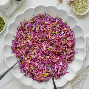 Large white scalloped bowl filled with shredded purple cabbage slaw with seeds, placed on a light surface with garlic cloves, spices, and utensils around. Two serving spoons rest in the bowl.