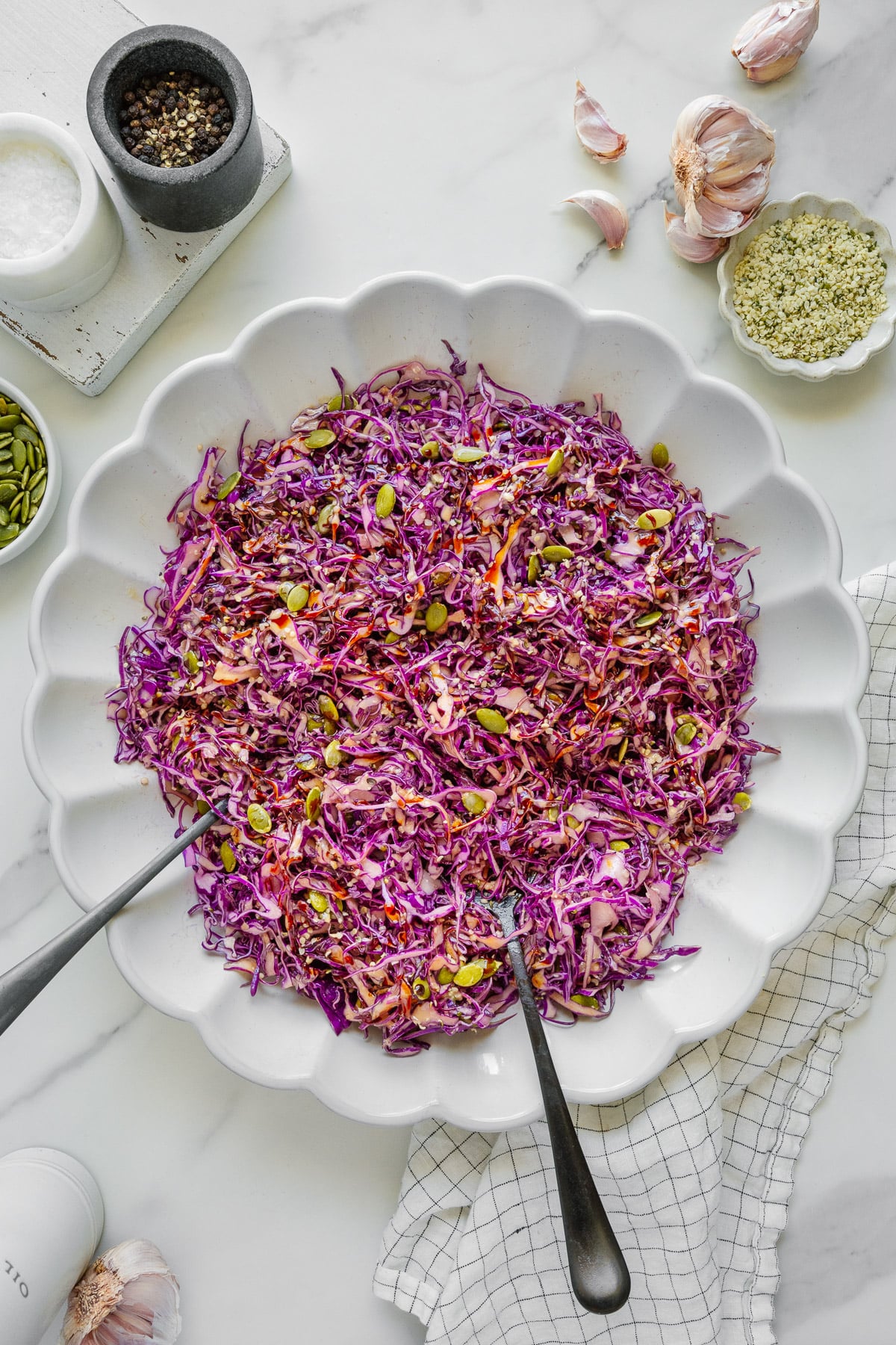 Large white scalloped bowl filled with shredded purple cabbage slaw with seeds, placed on a light surface with garlic cloves, spices, and utensils around. Two serving spoons rest in the bowl.
