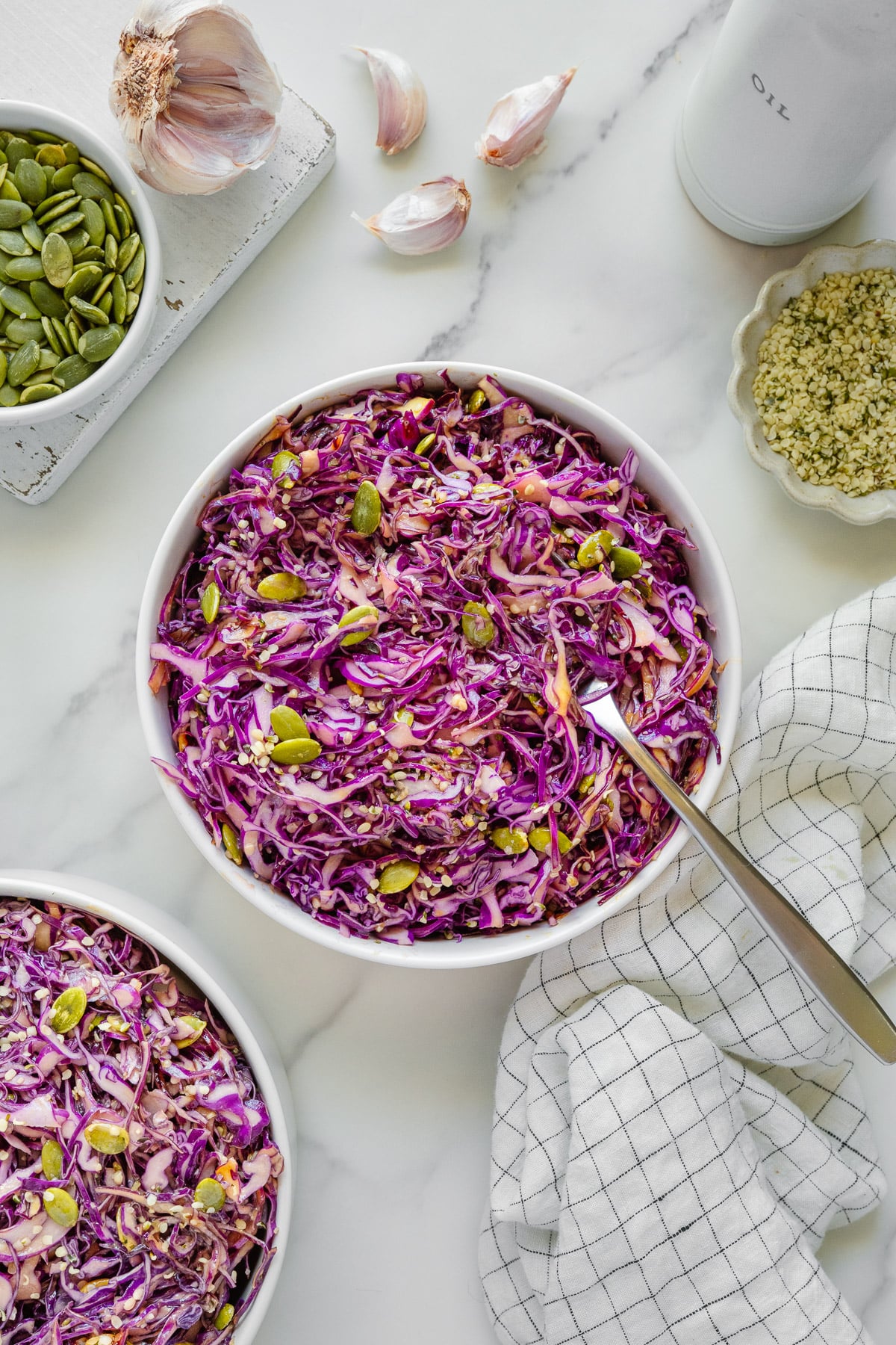 A white bowl filled with purple cabbage salad, topped with pumpkin seeds and hemp seeds. Garlic, a dish of seeds, and a bottle of oil are nearby on a marble surface. A checkered cloth is beside the bowl.
