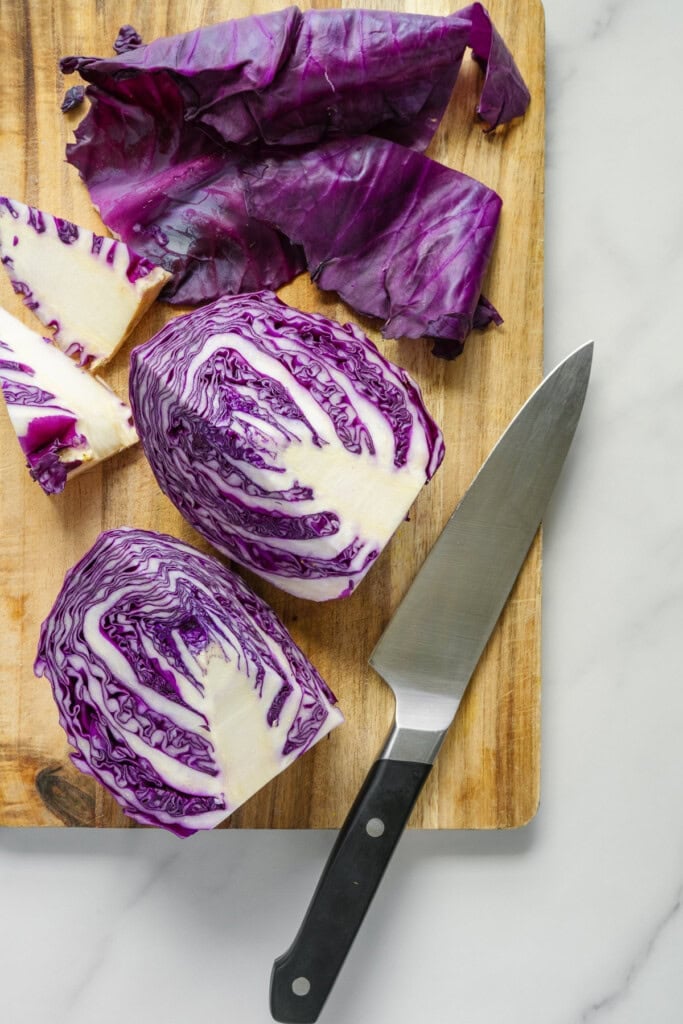 Sliced red cabbage and loose leaves on a wooden cutting board, with a kitchen knife nearby, all on a white marble surface.
