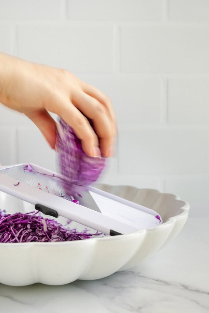 A hand slicing purple cabbage with a mandoline slicer over a white scalloped bowl, with shredded cabbage collecting in the bowl. White tiled background.