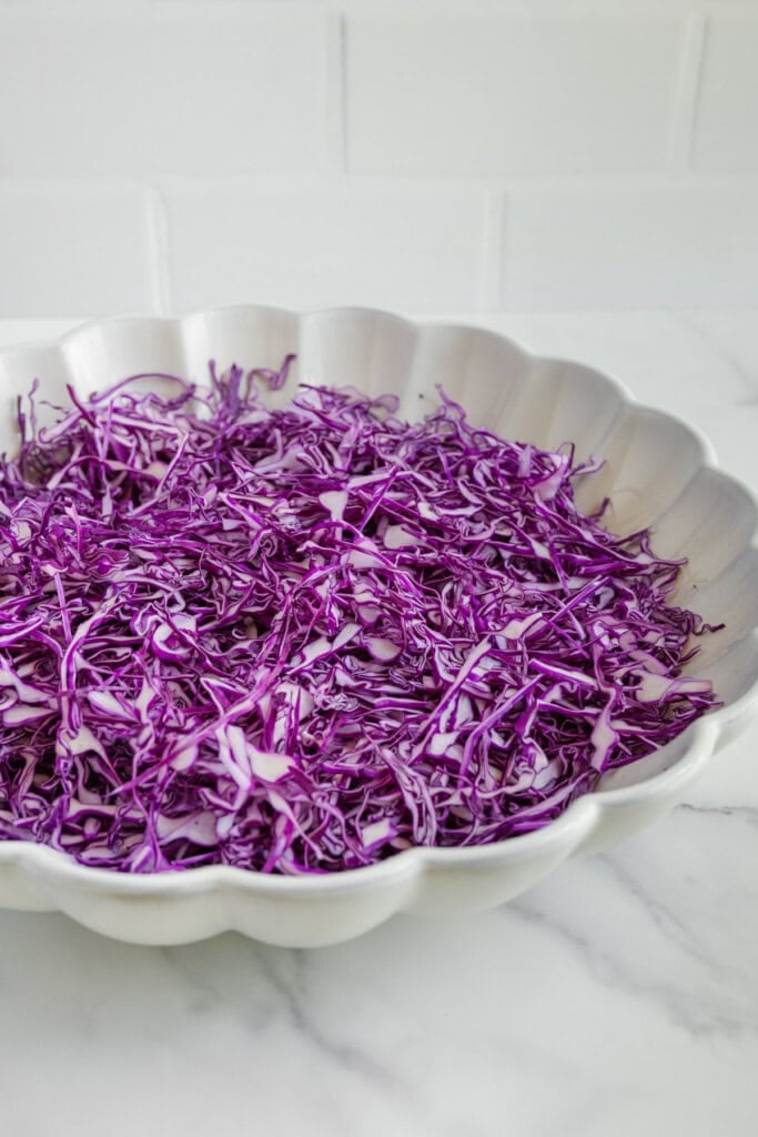 A scalloped white bowl filled with thinly shredded purple cabbage sits on a white marble countertop with a white tiled background.