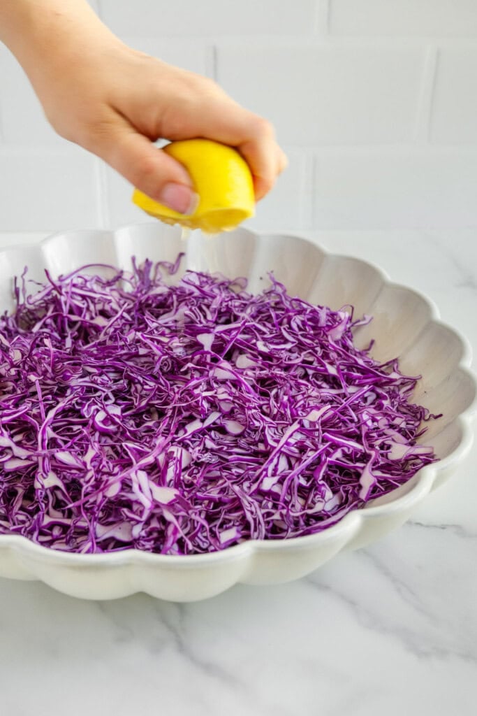 A hand squeezes a lemon over a white scalloped bowl filled with thinly sliced purple cabbage on a marble countertop.