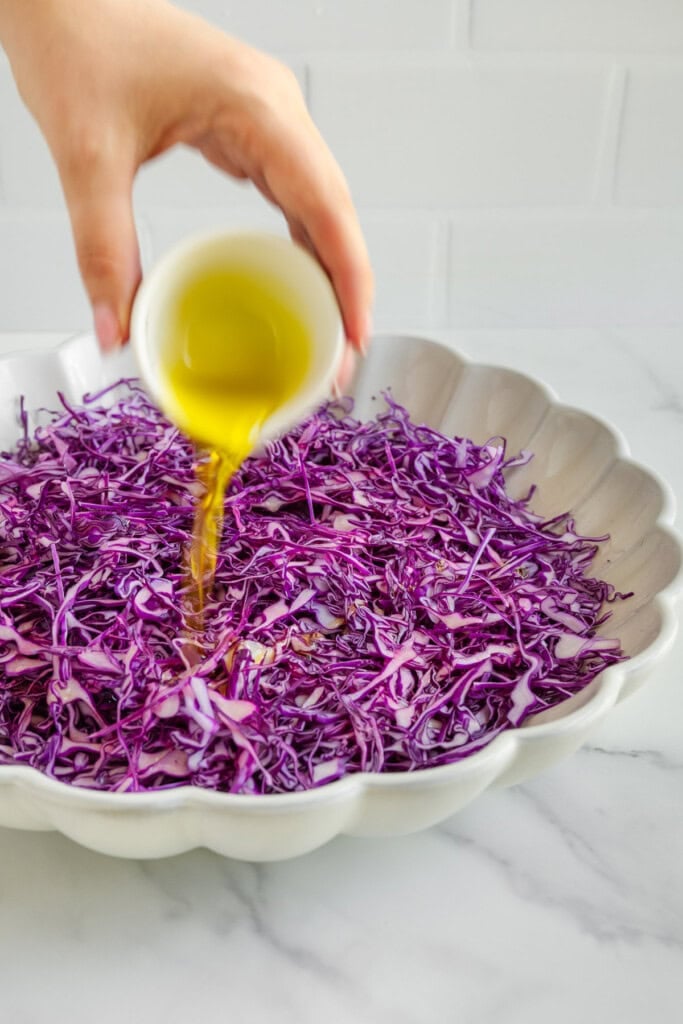 A hand pours olive oil from a small white cup over finely shredded purple cabbage in a large scalloped bowl on a marble countertop.