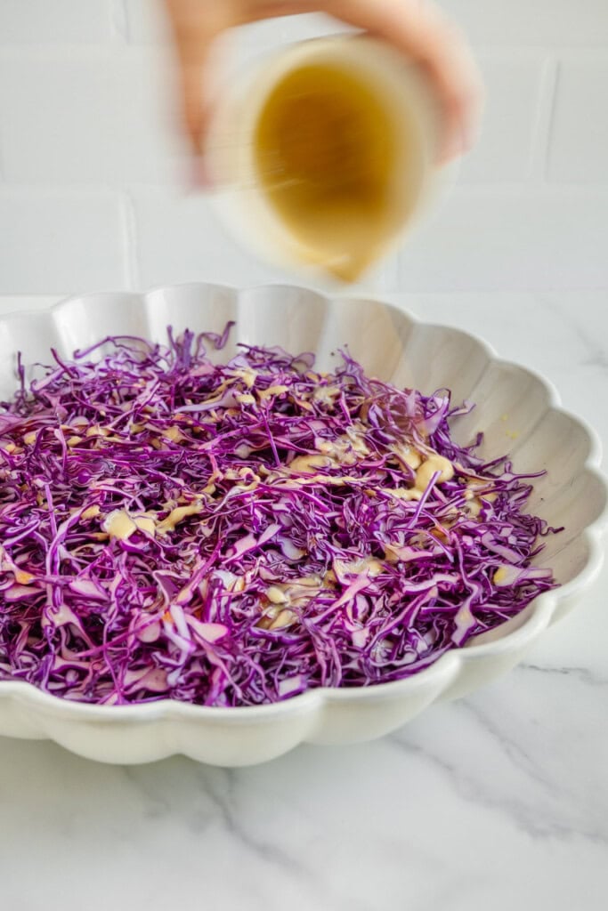 A hand pours dressing onto shredded purple cabbage in a scalloped white bowl, set on a marble surface with a white tiled background.