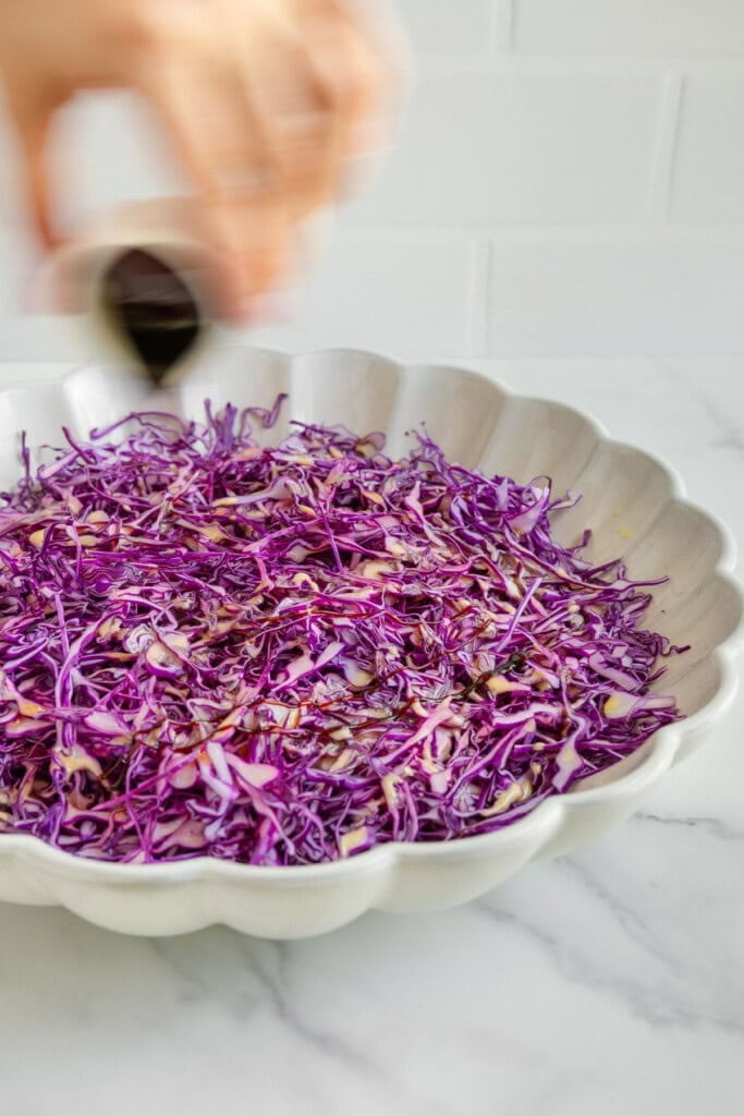 A hand pours dark liquid over shredded purple cabbage in a white scalloped bowl on a marble surface, with a white tile background.