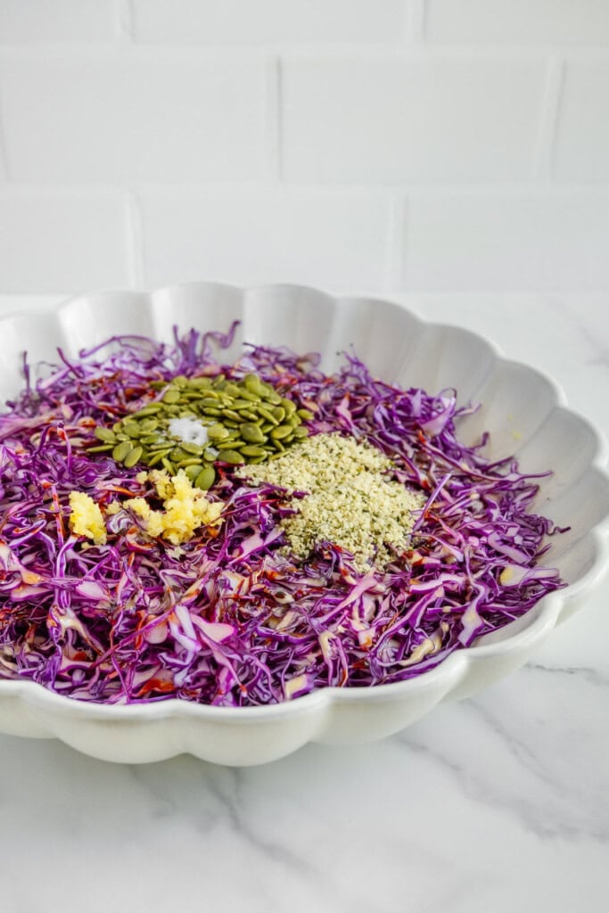 A white scalloped bowl contains shredded purple cabbage, grated ginger, pumpkin seeds, and hemp seeds, placed on a marble countertop with a white tiled background.