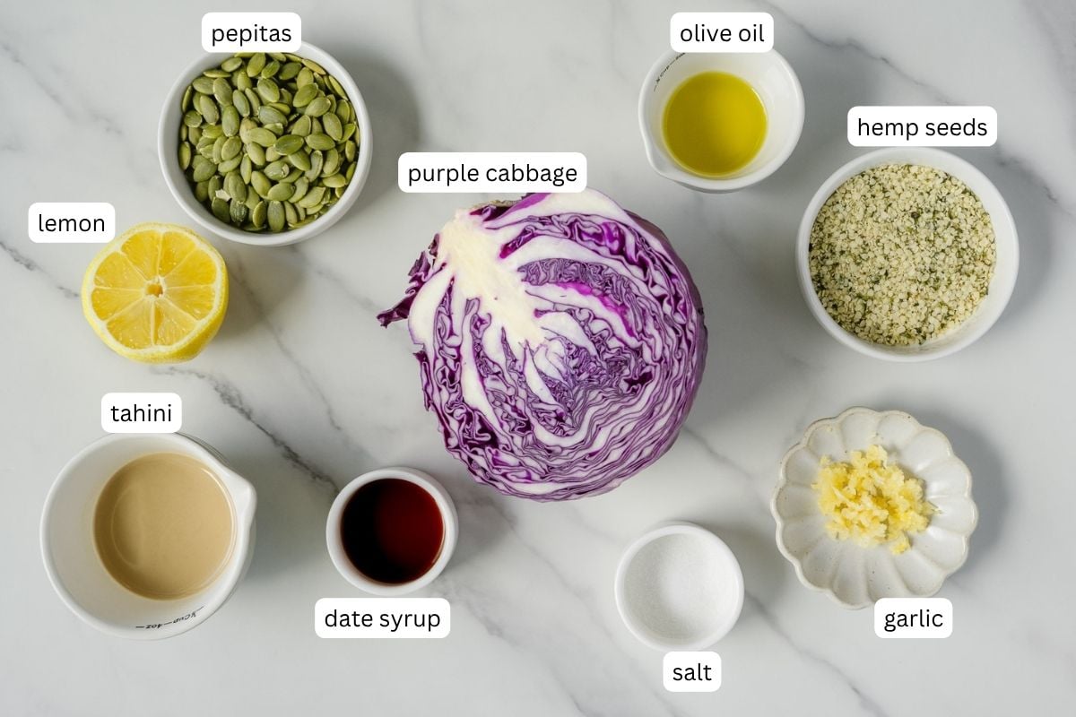 Ingredients for a healthy coleslaw or dressing are arranged on a marble surface: half a purple cabbage, lemon, tahini, date syrup, pepitas, olive oil, hemp seeds, salt, and minced garlic, each labeled.