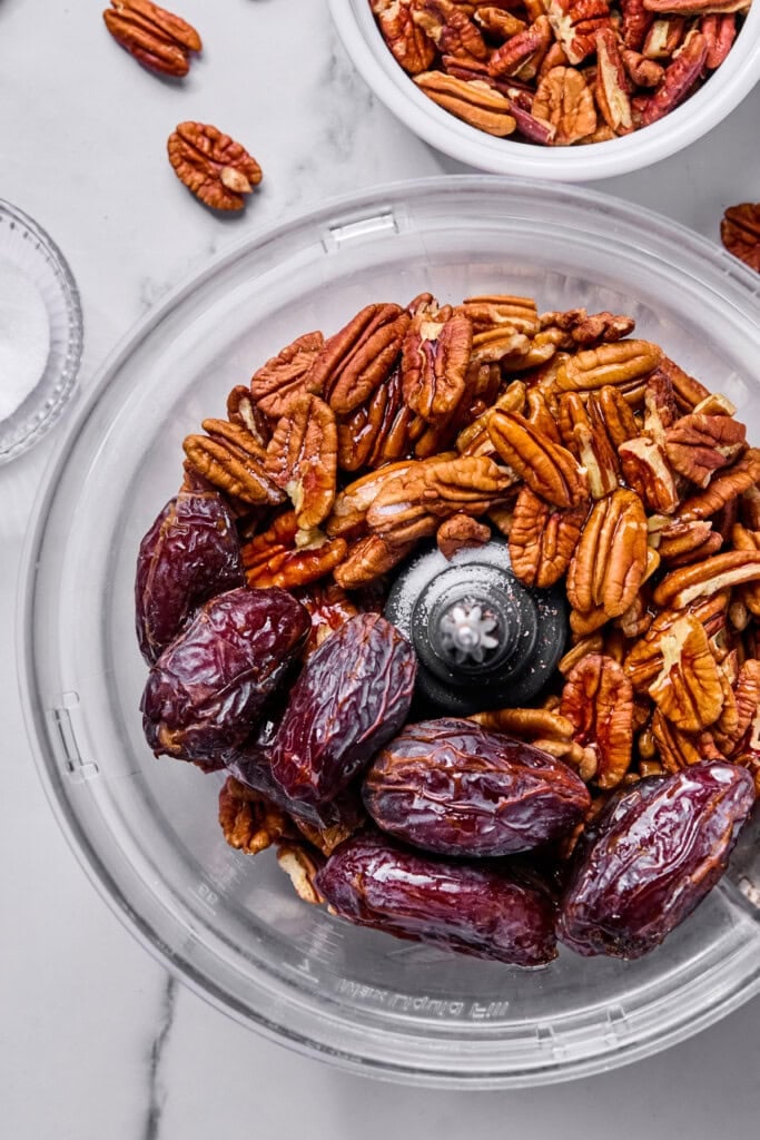A food processor bowl filled with whole pecans and large pitted dates, ready to be blended. Nearby are a small bowl of pecans and a small dish of salt on a marble countertop.