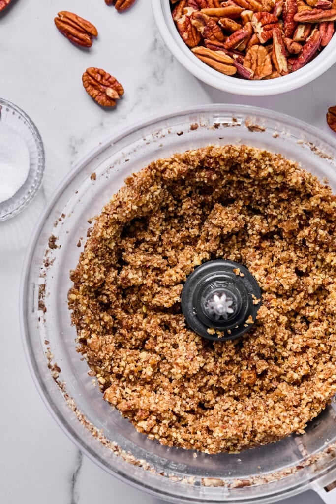 A food processor filled with blended pecans sits on a marble counter. Nearby are a small bowl of whole pecans, a few loose pecans, and a small dish of sugar.