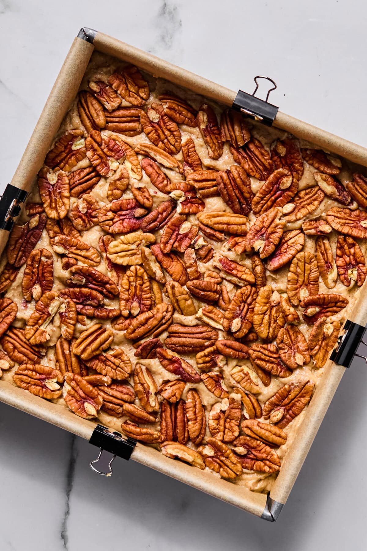 A square baking pan lined with parchment paper holds a layer of pecan halves arranged on top of a baked dessert, possibly pecan bars. Black binder clips secure the parchment to the sides of the pan.