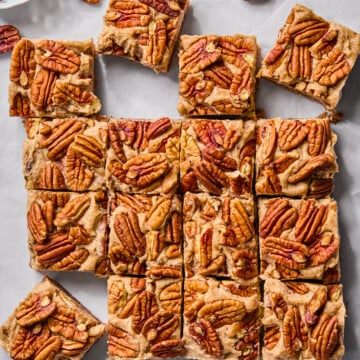 Sixteen pecan-topped dessert bars are arranged on parchment paper, with a few whole pecans scattered around on a marble surface.