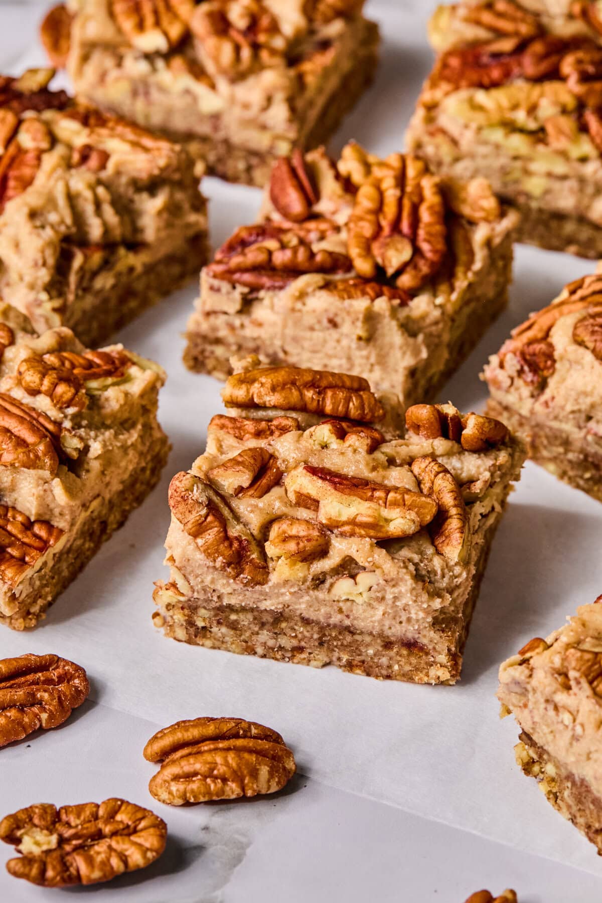 Nine pecan-topped dessert bars are arranged on white parchment paper, with several shelled pecans scattered around. The bars have a soft texture and a layer of whole pecans on top.