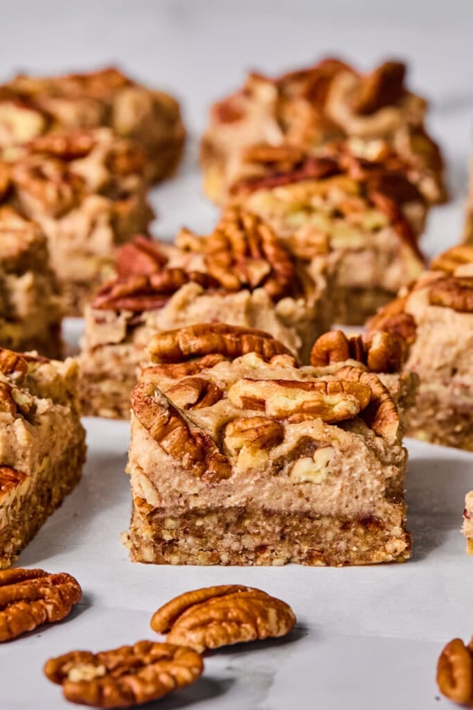 Close-up of pecan bars topped with whole pecans, arranged on a white surface with extra pecan halves scattered around. The bars have a creamy, textured layer and a nutty, crumbly base.