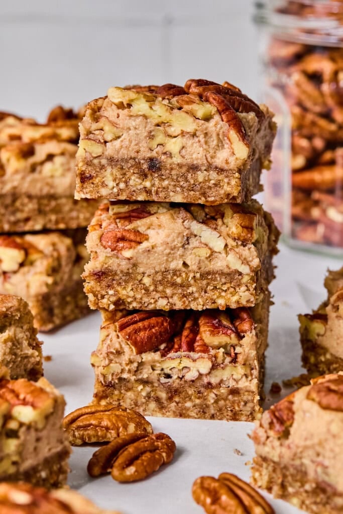 Three pecan bars stacked on top of each other, surrounded by more pecan bars and whole pecans, with a glass jar filled with pecans in the background.