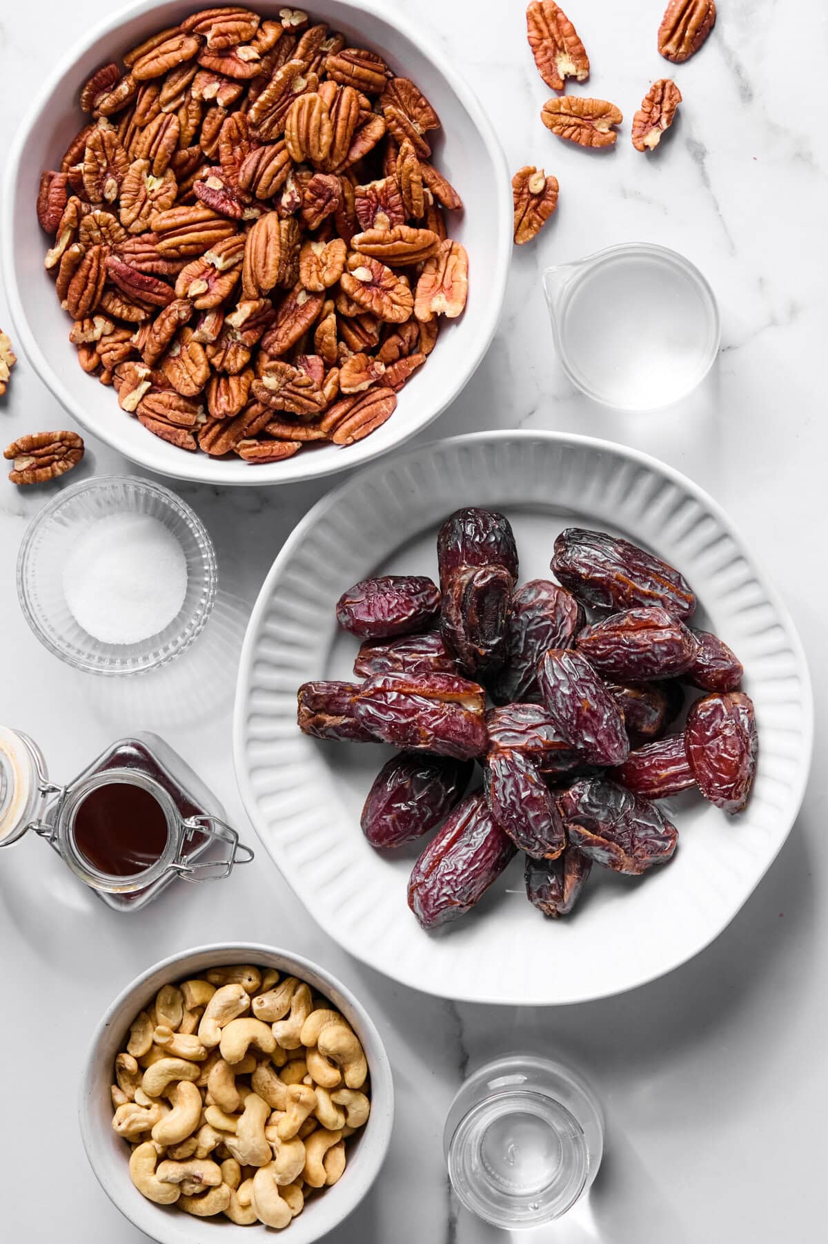 Bowl of pecans, bowl of dates, bowl of cashews, small glass bowls of sugar, water, and vanilla on a white marble surface. Some scattered pecans around. Overhead view.