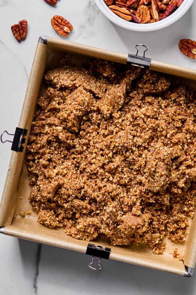 A baking pan lined with parchment paper holds a crumbly oat and pecan mixture. A bowl of pecans and a few loose pecans are nearby on a marble surface.