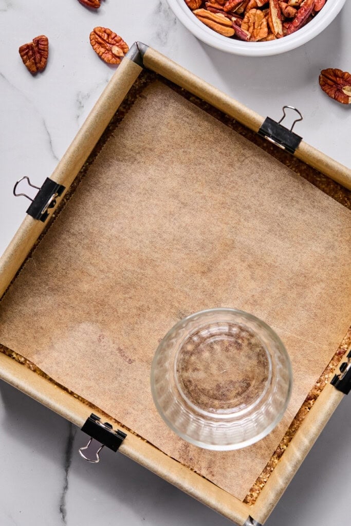 A square baking pan lined with parchment paper and clips holds a flat layer. A drinking glass sits on top. Nearby are pecans, both loose and in a white bowl, on a marble surface.