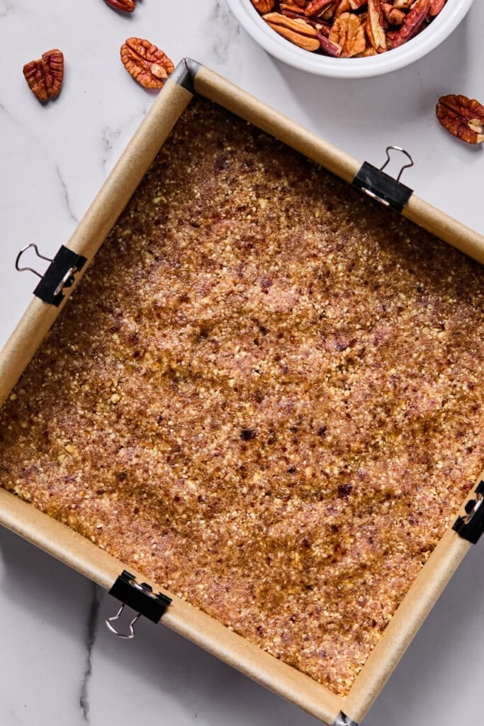 A parchment-lined square pan with a pressed crust mixture, clipped at the sides, sits on a marble surface near scattered pecans and a bowl of pecan halves.