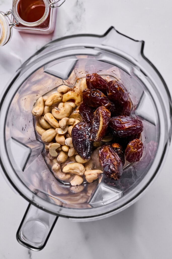 A blender filled with cashews, pitted dates, a splash of liquid, and a small amount of white powder, viewed from above on a white surface. A jar of dark syrup is partially visible nearby.