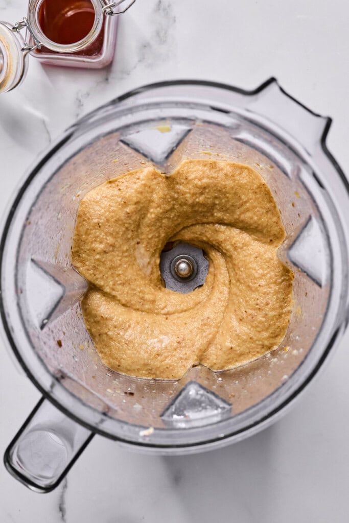 A blender with creamy, light brown mixture inside, viewed from above. Two small jars, one with amber liquid, are near the blender on a white surface.