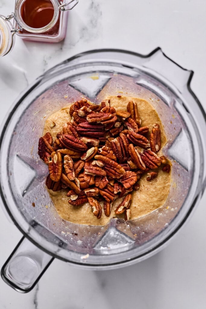 A blender filled with a thick, tan batter topped with whole pecans, viewed from above. Nearby are small jars containing honey or syrup and a light-colored liquid.