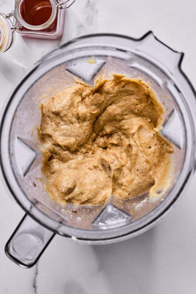 A blender filled with thick, creamy tan-colored batter sits on a white surface. Nearby are small glass jars, one containing a reddish liquid and the other with a yellowish substance.