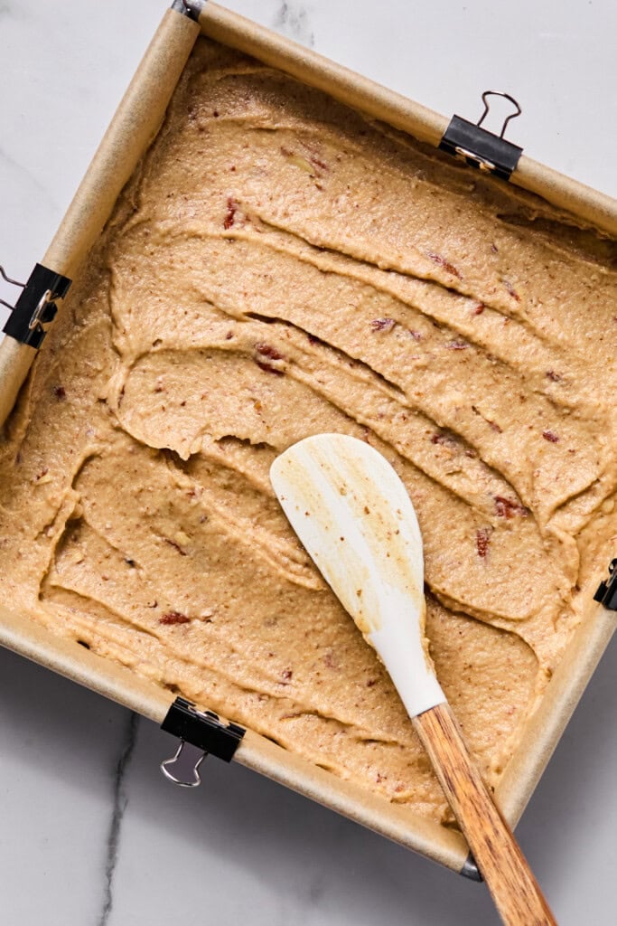 A square baking pan lined with parchment paper is filled with a thick, light brown batter. A white spatula with a wooden handle rests on top, and binder clips hold the paper in place.